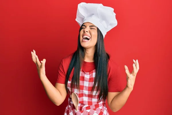 パン屋の制服を着た若い美しいヒスパニック系の女の子は 積極的な表現と腕を上げて狂った叫びと叫んで帽子を調理します フラストレーションコンセプト — ストック写真
