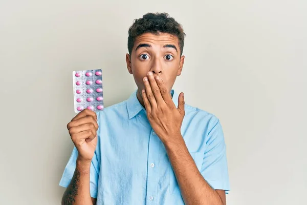 若いハンサムなアフリカ系アメリカ人の男性が手で口をカバー錠剤を保持し ショックを受け ミスを恐れている 驚きの表情 — ストック写真