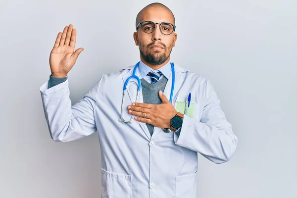 Hispanic adult man wearing doctor uniform and stethoscope swearing with hand on chest and open palm, making a loyalty promise oath