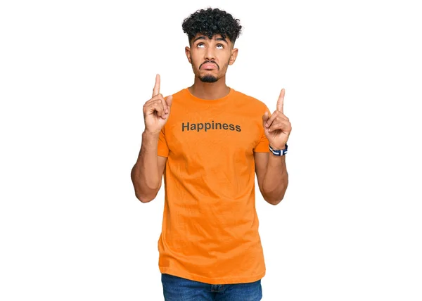年轻的阿拉伯男子穿着印有快乐字样的T恤衫 面带微笑地抬起头 用手指指示方向 忧郁而不快乐 — 图库照片
