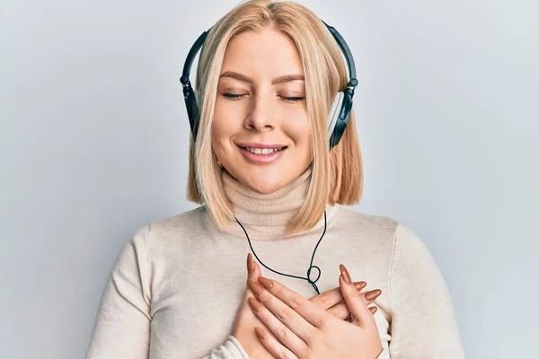 目を閉じて胸の上で手で微笑むヘッドフォンを使用して音楽を聴く若いブロンドの女性と顔に感謝のジェスチャー 健康の概念 — ストック写真