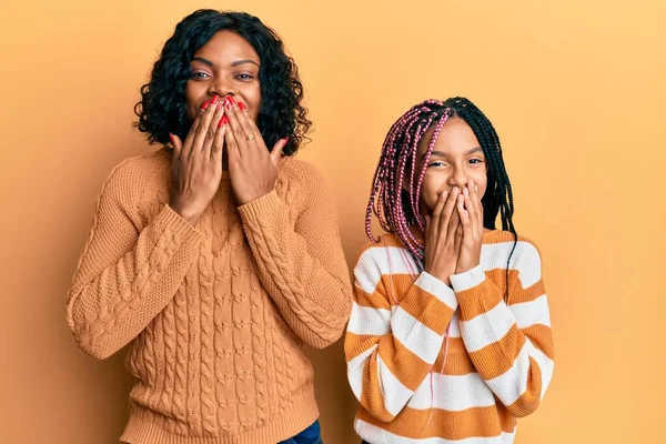 Beautiful african american mother and daughter wearing wool winter sweater laughing and embarrassed giggle covering mouth with hands, gossip and scandal concept