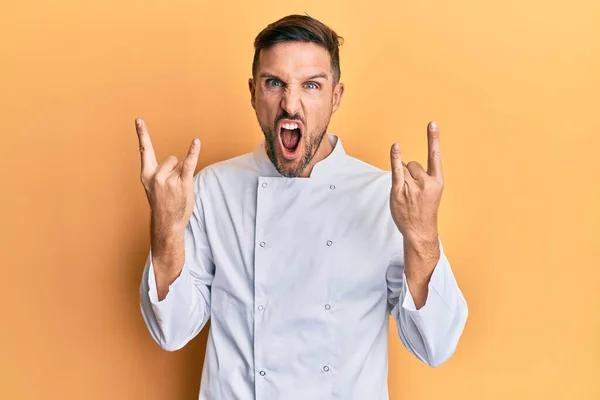 Handsome man with beard wearing professional cook uniform shouting with crazy expression doing rock symbol with hands up. music star. heavy concept.