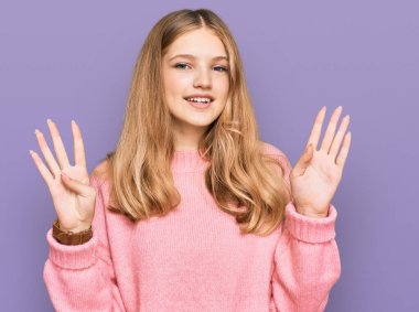 Güzel, genç, beyaz bir kız, günlük kışlık süveter giyiyor ve dokuz numaralı parmaklarıyla işaret ediyor. Kendinden emin ve mutlu bir şekilde gülümsüyor.. 