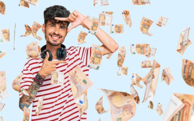 Genç İspanyol adam müzik dinliyor kulaklık takıyor gülümsüyor ve elleri ve parmakları gülen yüzüyle çerçeve yapıyor. yaratıcılık ve fotoğrafçılık kavramı.