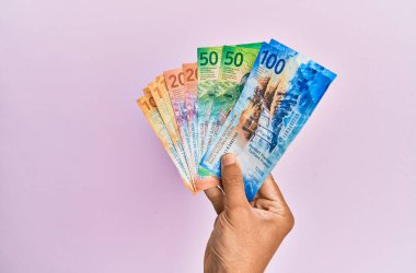 İspanyol eli İsviçre frangı banknotlarını soyutlanmış pembe arka planda tutuyor.