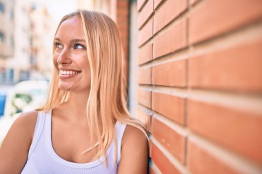 Genç sarışın kız mutlu bir şekilde gülümsüyor. Şehrin duvarına yaslanıyor..