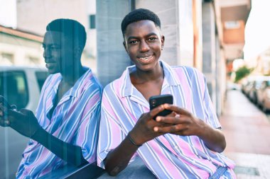 Afro-Amerikalı genç adam mutlu gülümsüyor. Şehrin caddesinde duvara yaslanmış akıllı telefon kullanıyor..