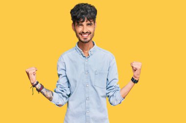 Gündelik kıyafetler giyen genç İspanyol adam çok mutlu ve heyecanlı. Kazanan jestler yapıyor. Kolları havada, gülümsüyor ve başarı için bağırıyor. kutlama konsepti. 