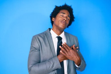 Afro saçlı, iş ceketi giyen, göğsünde elleri göğsünde gülümseyen, yüzünde minnet ifadesi olan yakışıklı Afro-Amerikan erkeği. sağlık kavramı. 