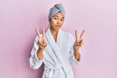 Duş havlusu şapkası ve bornoz giymiş genç Çinli kadın kameraya bakıp zafer işareti yapıyor. İki numara.. 