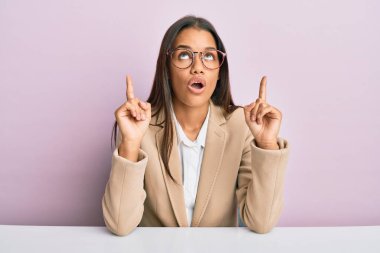 Ofiste çalışan güzel İspanyol kadın şaşkın şaşkın yukarı bakıyor ve parmaklarıyla işaret ediyor ve kollarını kaldırıyor.. 