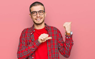 İspanyol asıllı, gündelik giysiler giyen, elleri ve başparmakları havada, kendinden emin bir şekilde arkasını gösteren genç bir adam. 