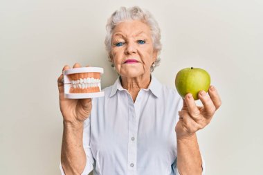 Senior grey-haired woman holding green apple and denture teeth relaxed with serious expression on face. simple and natural looking at the camera.  clipart