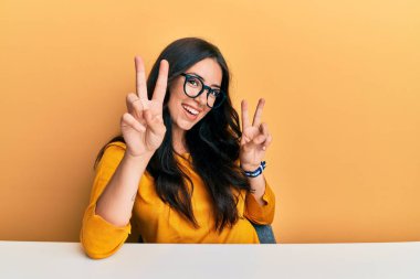 Güzel esmer, gözlüklü, sıradan giysiler giyen, masaya oturmuş gülümseyen ve parmaklarını zafer işareti yapan genç bir kadın. İki numara.. 