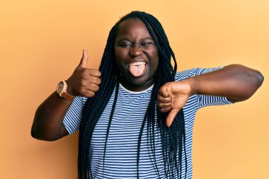 Örgülü genç siyahi kadın baş parmağını kaldırıyor ve parmağını kaldırıyor. Komik bir ifadeyle dilini dışarı çıkarıyor.. 