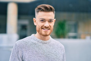 Genç beyaz adam gülümseyerek şehirde mutlu mesut duruyor..