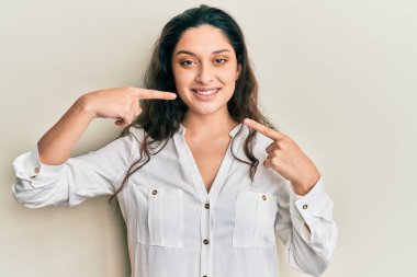 Güzel bir ortadoğulu kadın günlük kıyafetler giyip neşeli bir şekilde gülümsüyor ve parmaklarıyla ve ağzıyla işaret ediyor. diş sağlığı kavramı. 