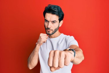 Latin kökenli genç adam, günlük beyaz tişört giyiyor dövüşmek için yumruk atıyor agresif ve öfkeli saldırı, tehdit ve şiddet. 