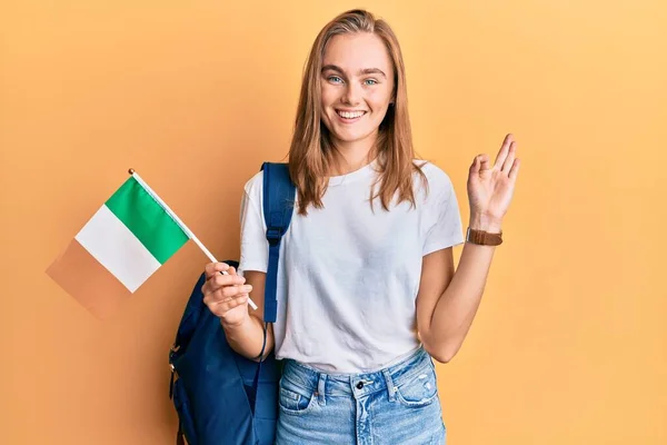 Beautiful blonde woman exchange student holding ireland flag doing ok sign with fingers, smiling friendly gesturing excellent symbol