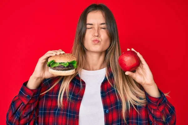 美丽的高加索女人比较汉堡包和健康的红苹果 看着镜头吹一个吻是可爱和性感的 爱的表达 — 图库照片