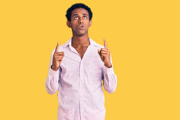 アフリカのハンサムな男を着てカジュアルなピンクのシャツ驚きと驚きを見上げと指差すと指と上げ腕 — ストック写真