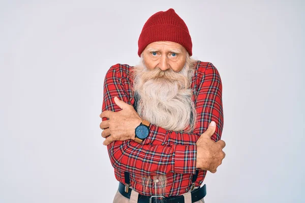 Old senior man with grey hair and long beard wearing hipster look with wool cap shaking and freezing for winter cold with sad and shock expression on face