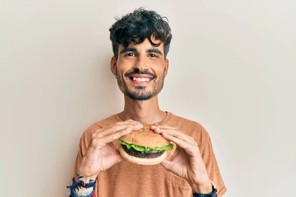 年轻的惊慌失措的男人吃着美味的经典汉堡 脸上挂着快乐而凉爽的笑容 显示牙齿 — 图库照片