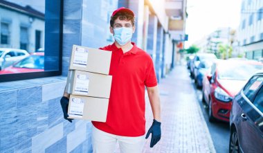 Coronavirus koruması takan beyaz bir kurye elinde karton paketler, şehirde yürüyor..