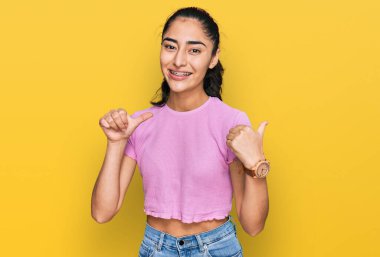 Diş teli takan, sıradan kıyafetler giyen İspanyol bir genç kız. Arka tarafı işaret ediyor. El ve baş parmakları havada, kendinden emin. 