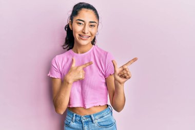 Diş teli takan, sıradan kıyafetler giyen, gülümseyen ve kameraya bakan iki eli ve parmakları yanda olan İspanyol bir genç kız.. 