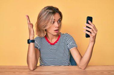 Beautiful caucasian woman doing video call waving to smartphone in shock face, looking skeptical and sarcastic, surprised with open mouth  clipart