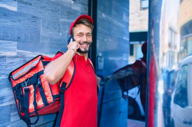 Beyaz teslimatçı, kırmızı üniformalı ve teslimat çantalı mutlu bir şekilde dışarıda telefonda konuşuyor.