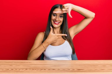 Latin kökenli genç bir kadın, gündelik giysiler giymiş masaya oturmuş gülümsüyor ve elleri ve parmakları mutlu yüzlü bir çerçeve yapıyor. yaratıcılık ve fotoğrafçılık kavramı. 