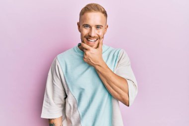 Gündelik kıyafetler giyen genç beyaz adam kameraya kendinden emin bakıyor kollarını kavuşturmuş ve çenesini kaldırmış gülümsüyor. Olumlu düşünmek. 