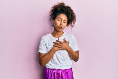 Afro saçlı, spor giyimli, göğsünde elleri kapalı, yüzünde minnet dolu bir jestle gülümseyen güzel bir Afrikalı Amerikalı kadın. sağlık kavramı. 