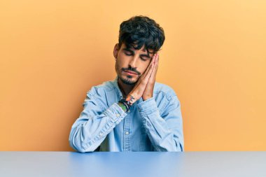 Latin asıllı genç adam, gündelik giysiler içinde masada oturuyor. Yorgun uyuyor, hayal kuruyor ve elleri ile poz veriyor. Gözleri kapalı gülümsüyor.. 