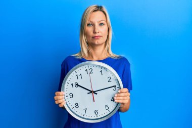 Güzel, orta yaşlı, sarışın bir kadın büyük bir saat tutuyor. Yüzünde ciddi bir ifade var. Basit ve doğal bir şekilde kameraya bakıyorsun.. 