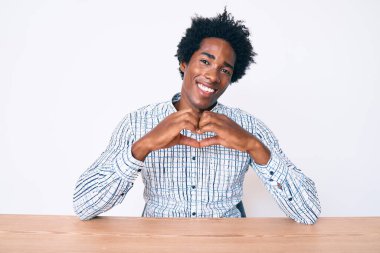 Yakışıklı Afro saçlı, sıradan kıyafetler giyen, aşık bir şekilde masasında oturan ve elleriyle kalp sembolü ve şekli gösteren Afro-Amerikan bir adam. romantik kavram. 