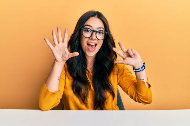 Güzel, esmer, gözlüklü ve günlük kıyafetler giyen genç bir kadın masanın üzerinde oturuyor ve sekiz numaralı parmaklarıyla işaret ediyor kendinden emin ve mutlu bir şekilde gülümsüyor.. 