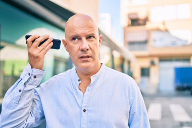 Şehirdeki akıllı telefonu kullanarak sesli mesaj dinleyen, ciddi ifadeli orta yaşlı kel bir adam..