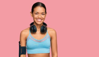 Spor kıyafetleri ve kulaklık takan güzel İspanyol kadın pozitif ve mutlu görünüyor ve kendine güvenen bir gülümsemeyle dişlerini gösteriyor. 