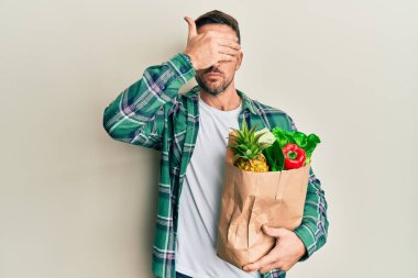 Sakallı yakışıklı bir adam elinde poşetle market poşetleriyle gözlerini kapamış, ciddi ve üzgün görünüyor. Görme, saklama ve reddetme konsepti 