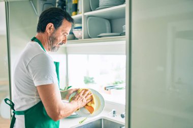Sakallı, orta yaşlı bir adam mutlu bir şekilde evde bulaşık yıkıyor.