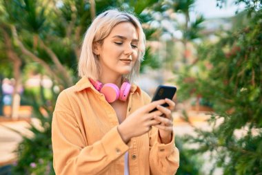 Parkta akıllı telefon ve kulaklık kullanarak gülümseyen genç sarışın kız..