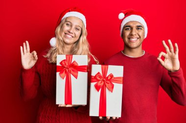 Irklar arası genç bir çift Noel şapkası takıyor ve ellerinde bir hediye var. Parmaklarıyla imza atıyor, gülümsüyor ve mükemmel bir sembol. 