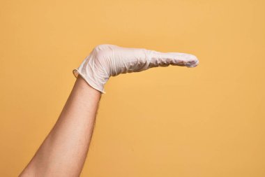 Tıbbi eldiveni olan, sarı arka planda, düz palmiye ürünü sunan, el hareketi yapan, boş fotokopi alanı sunan beyaz bir gencin eli.