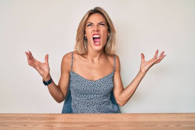 Kafkasyalı güzel bir kadın, gündelik giysiler içinde masada oturmuş deli deli deli bağırıyor ve agresif bir ifadeyle bağırıyor ve kollarını kaldırıyor. hüsran kavramı. 