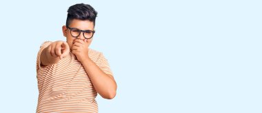 Günlük kıyafetler giyen ve gözlüklü küçük çocuk sana gülüyor, eli ağzında kamerayı işaret ediyor, utanç ifadesi var. 