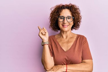 Güzel orta yaşlı yetişkin bir kadın, günlük kıyafetler ve gözlük takıyor, gülümsüyor, mutlu bir şekilde elini ve parmağını yana uzatıyor. 